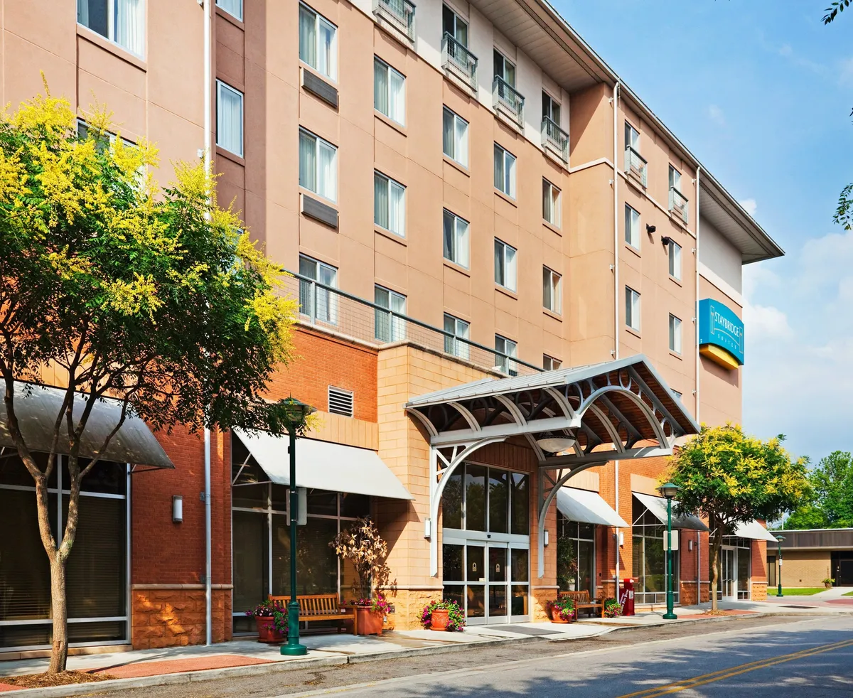 This exterior shot focuses on the inviting front entrance, with potted plants and large windows creating a bright, cheerful atmosphere. Guests are seen relaxing outside or checking in, reinforcing the sense of comfort and hospitality.