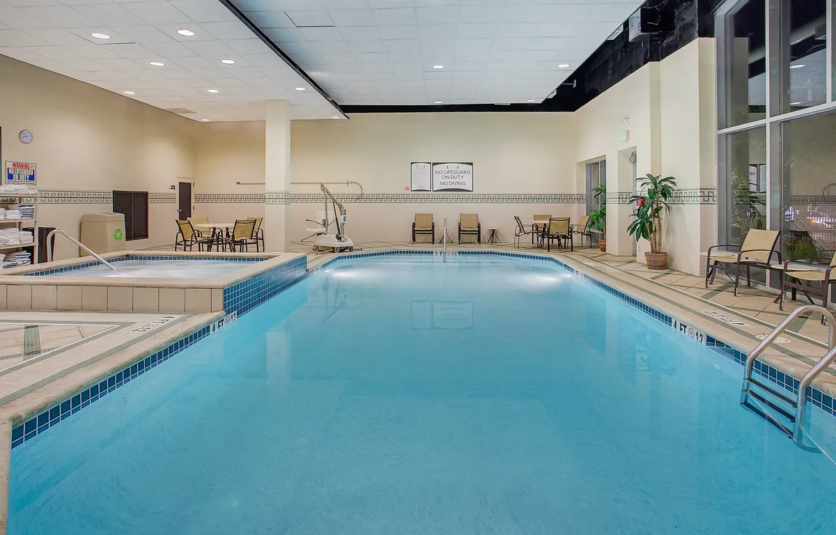 Inside, the tranquil indoor pool area features a swimming pool, jacuzzi, and comfortable lounge chairs surrounded by lush indoor plants. The space is designed for relaxation after a long day, with a peaceful ambiance enhanced by soft lighting and a visible clock.