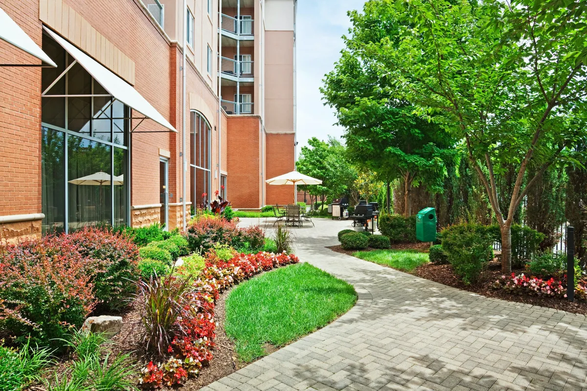 A bright, inviting exterior scene featuring lush flower beds, a neatly paved pathway leading to an outdoor seating area, and green trees surrounding the property. The space feels open and welcoming, ideal for families preparing for a day of city exploration or winding down after adventures.
