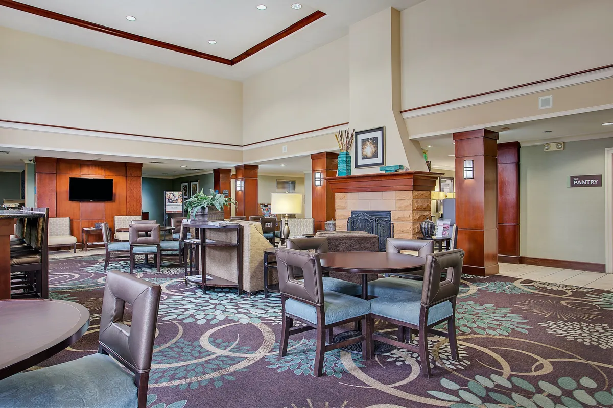 A cozy living area centered around a fireplace, with comfortable seating, decorative plants, and a pantry entrance visible in the background. The scene conveys warmth and a sense of home, ideal for relaxing, socializing, or enjoying entertainment together.