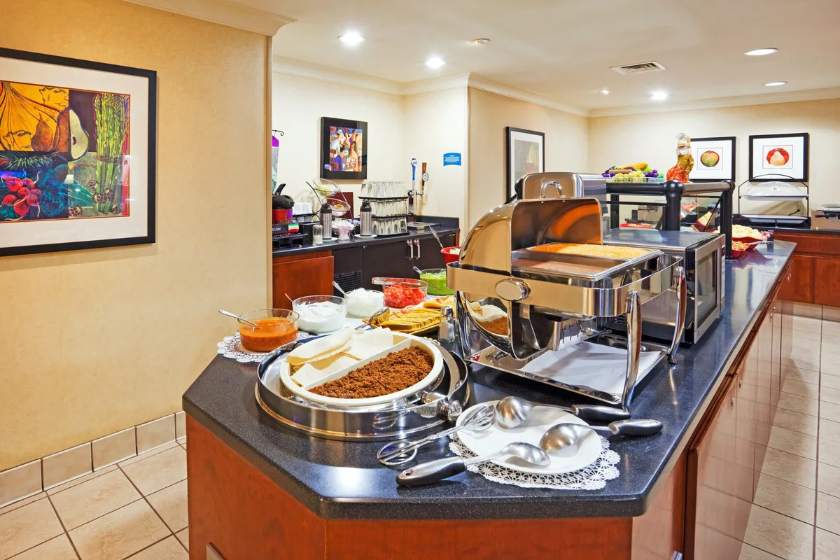 This interior scene features a lively buffet counter with a variety of food items and a coffee station, surrounded by decorative artwork. Guests gather for conversation and dining, evoking the convivial mood of evening guest receptions and shared meals.