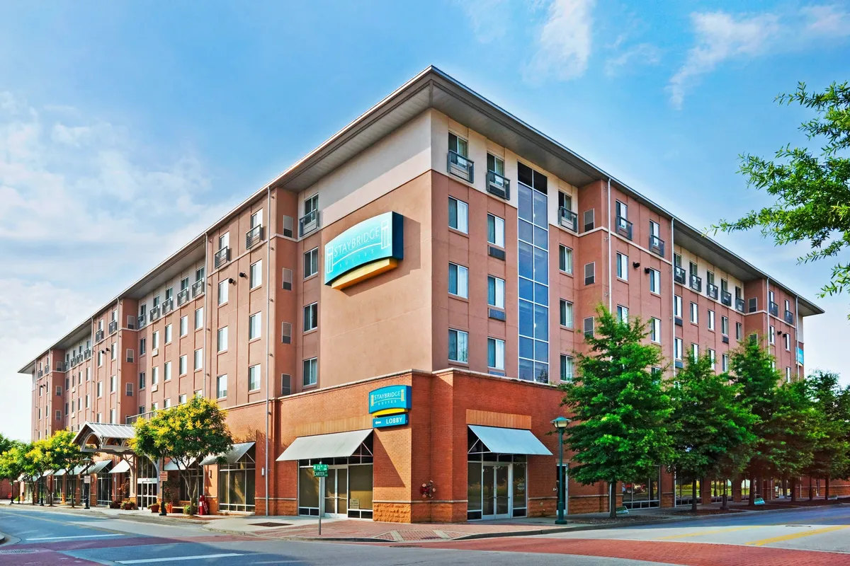 The image highlights the hotel's exterior facade, featuring a welcoming entrance awning, clear hotel signage, and mature trees that frame the building. In the scene, guests are seen checking in and enjoying the outdoor seating area, with the vibrant neighborhood as a backdrop.