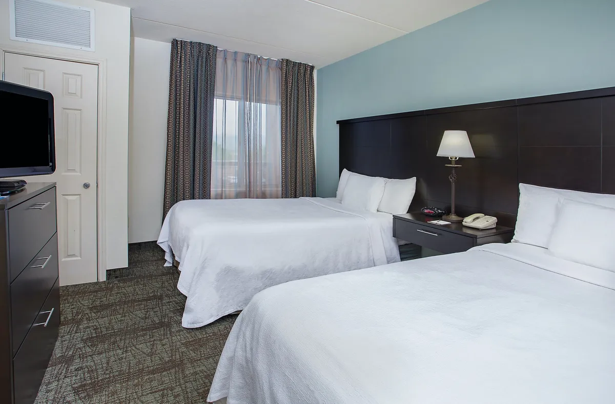A comfortable guest room featuring two double beds positioned beneath soft ambient lighting, with a nightstand and lamp between them. The room also includes a television, large windows draped with curtains, and inviting decor that signals a restful environment. The beds are made up with fresh linens, adding to the clean and serene atmosphere.