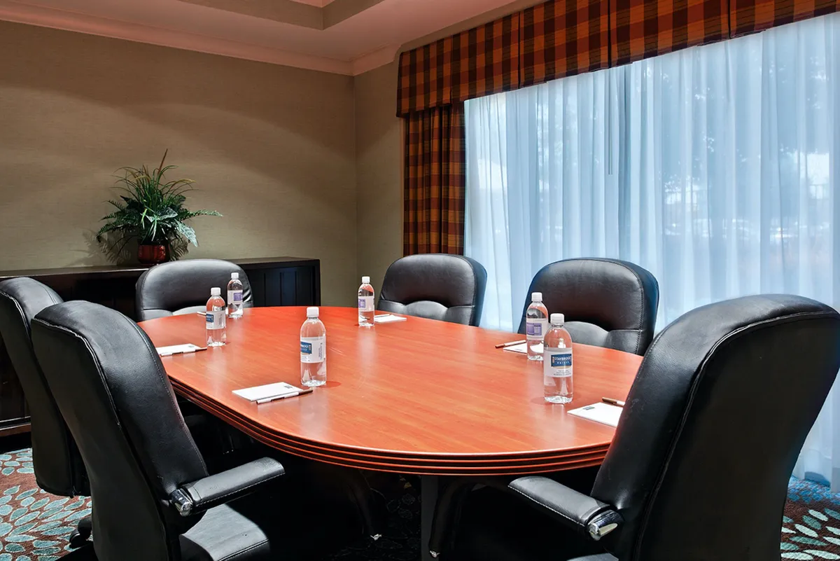 This interior shot highlights a modern meeting room with an oval wooden table, comfortable black leather chairs, water bottles neatly arranged, and a touch of greenery from a corner plant. Soft light streams in through sheer curtains, creating a professional yet welcoming space.