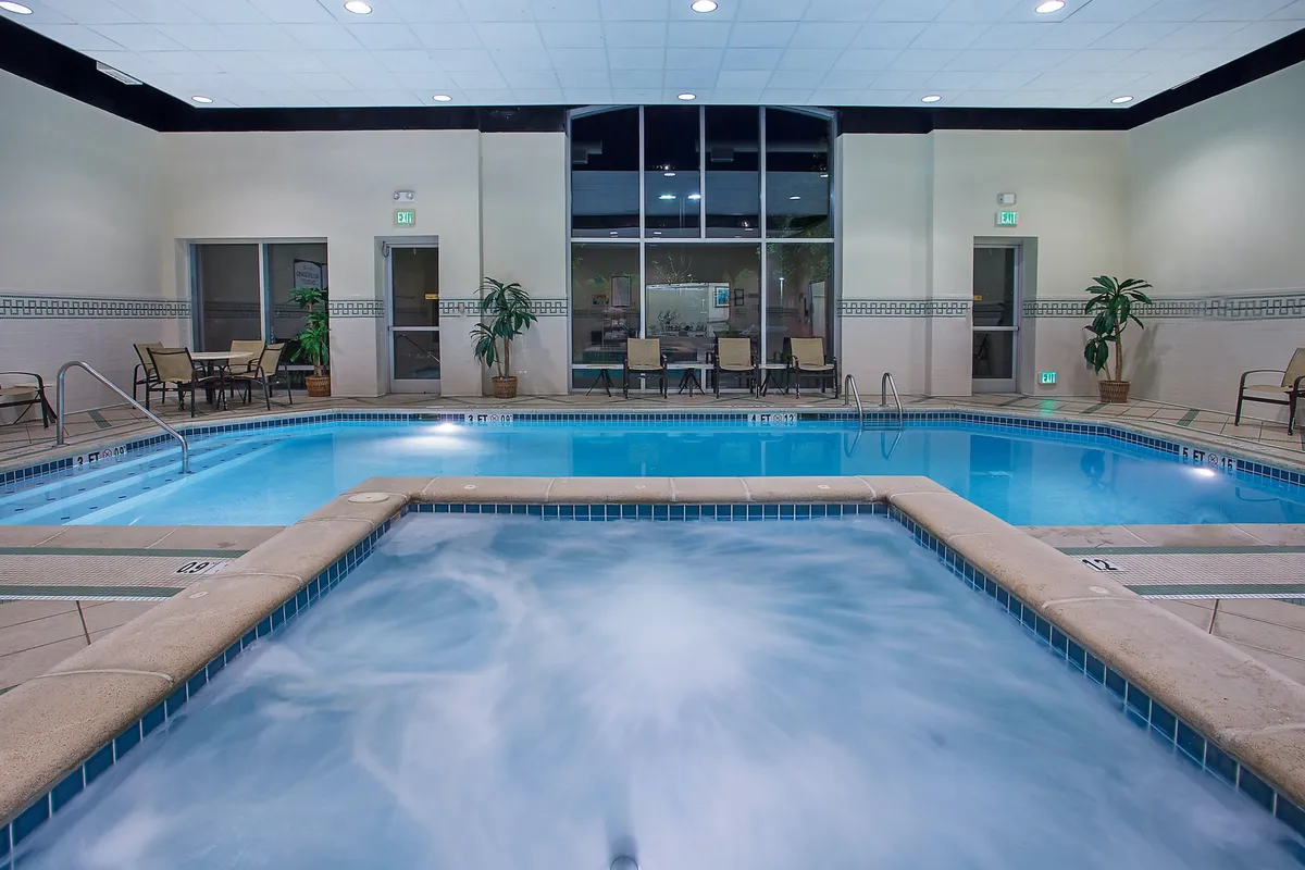 This interior image captures the hotel's inviting pool area, featuring a sparkling indoor pool surrounded by lush plants, a hot tub under soft, ambient lighting, and comfortable seating areas. Guests are seen swimming, relaxing in the hot tub, and socializing by the water, creating an atmosphere of tranquility and connection.