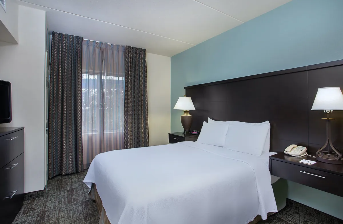 A tranquil bedroom space featuring a plush queen bed, flanked by lamps and framed by elegant curtains. The setting is serene, with a guest enjoying a book in bed or relaxing by the window with coffee, highlighting both relaxation and personal time. The room exudes privacy and calm, ideal for couples or solo travelers.
